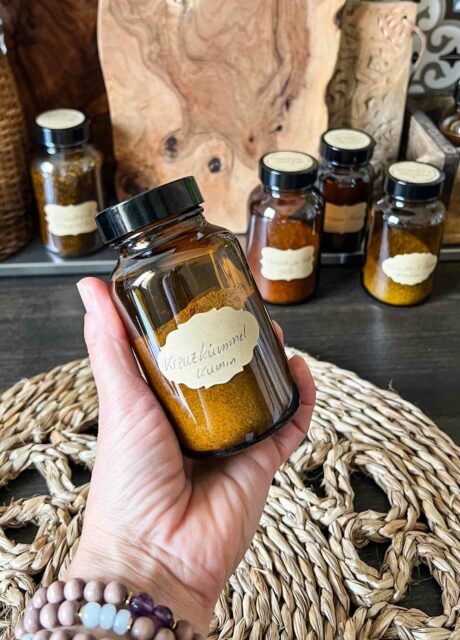Apothekerglas mit Kreuzkümmel und handbeschriftetem Kraft-Aufkleber in einer Hand, im Hintergrund weitere Gewürzgläser.