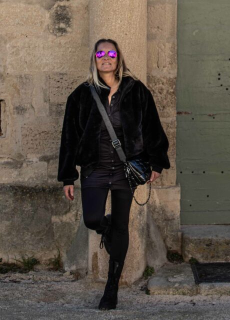 Frau lehnt lässig an einer Säule und trägt schwarze Leggings von Stronger Label. Sie kombiniert die Leggings mit einer schwarzen Jacke, Stiefeln, Sonnenbrille und einer schwarzen Umhängetasche.