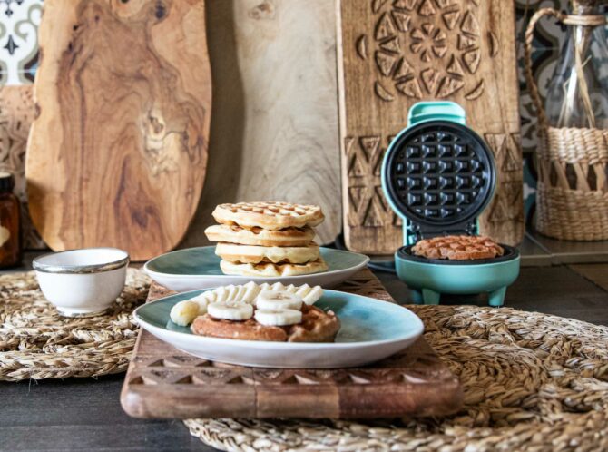 Stapel von frischgebackenen Waffeln, serviert auf einem rustikal gedeckten Tisch, mit Waffeleisen im Hintergrund und einer Waffel mit Bananenscheiben im Vordergrund