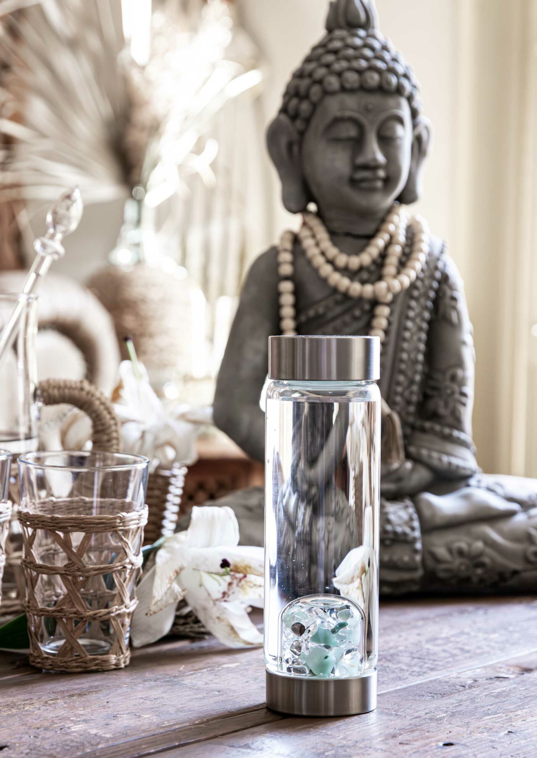 VitaJuwel ViA Edelstein Wasserflasche mit Aquamarin, Aventurin, Rauchquarz und Bergkristall auf einem Boho-Style Holztisch vor einer Buddha-Statue